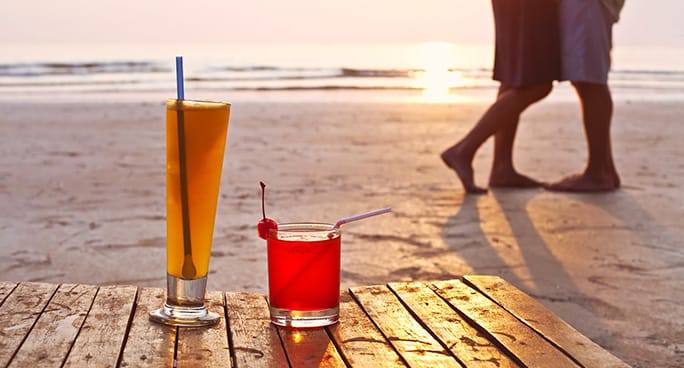 Due bicchieri di cocktail appoggiati a terra in riva al mare col tramonto in sottofondo