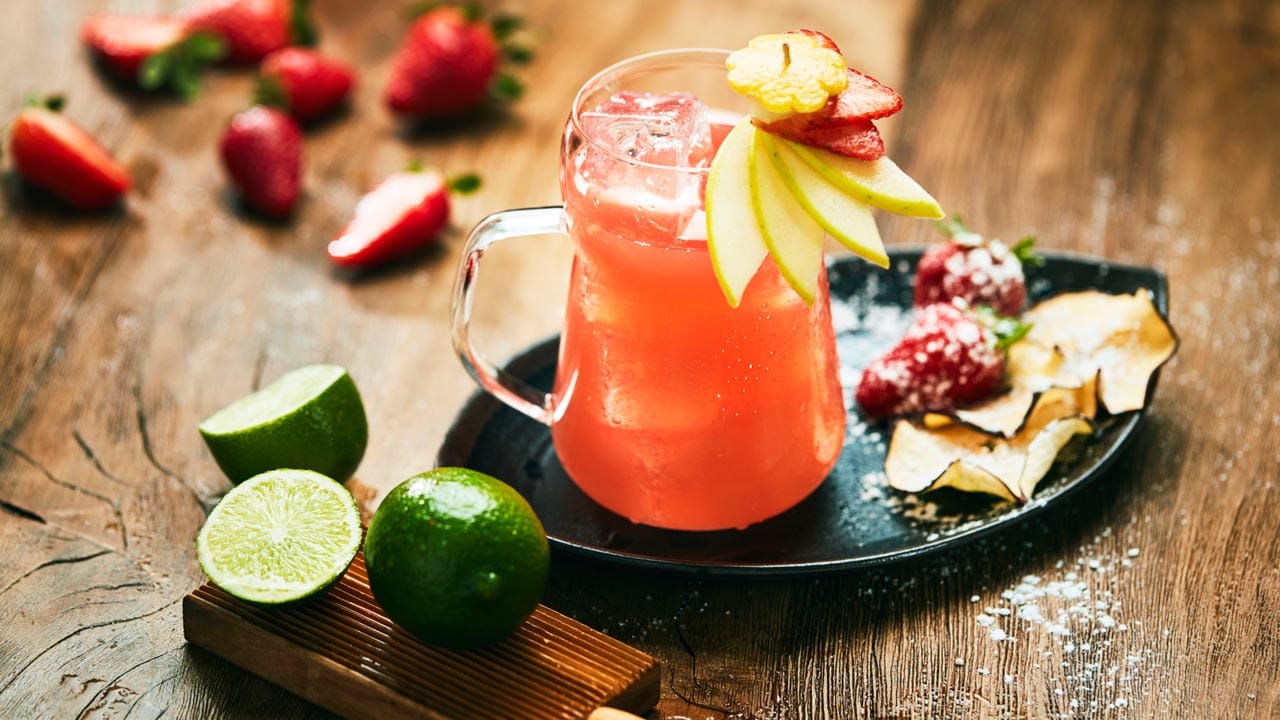 Il cocktail Emozioni in tazza decorato con fette di mela, contornato da lime e fragole